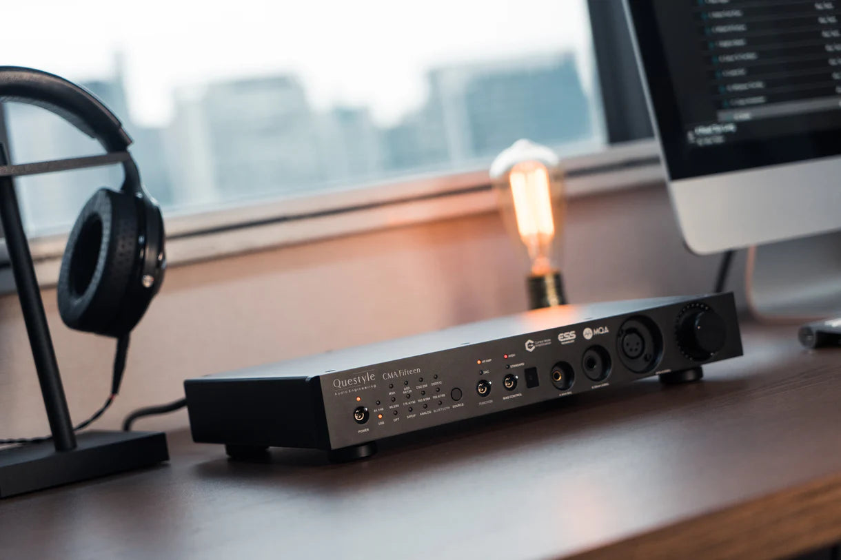 Questyle CMA Fifteen desktop DAC and headphone amplifier on a desk with headphones, lamp, and computer monitor in background