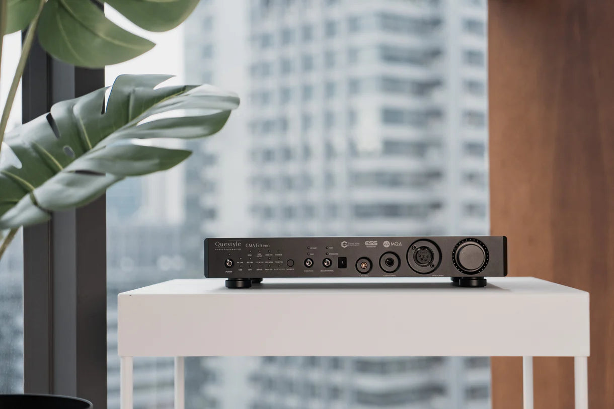 Questyle CMA Fifteen desktop DAC and headphone amplifier placed on a white table with cityscape background and green plant leaves